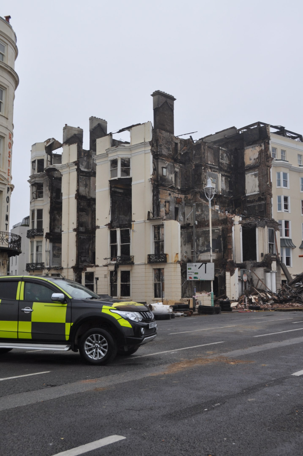 Ein Polizeiwagen vor einem stark beschädigten Gebäude mit zersplitterten Fenstern und verstreuter Trümmer, mit zusätzlichen Gebäuden und Himmel im Hintergrund.