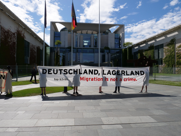 Gruppe von Menschen mit einem Transparent mit der Aufschrift "Deutschland, Lagerland Migration ist kein Verbrechen" vor einem eingezäunten Gebäude mit Bäumen und Flaggen.