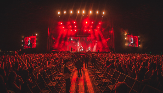 Eine große Menschenmenge steht vor einer hell beleuchteten Bühne mit Geländern, auf der Musiker spielen und andere zuschauen, vor einem dunklen Hintergrund.