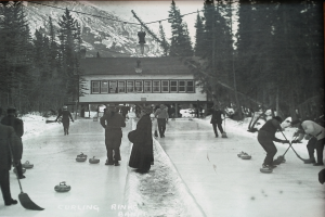 Eine Gruppe von Menschen, die Curling auf einer Eisbahn spielen, umgeben von Bäumen und einem Gebäude im Hintergrund, dargestellt in Schwarz-Weiß.