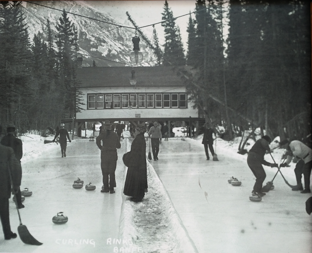 Eine Gruppe von Menschen, die Curling auf einer Eisbahn spielen, umgeben von Bäumen und einem Gebäude im Hintergrund, dargestellt in Schwarz-Weiß.