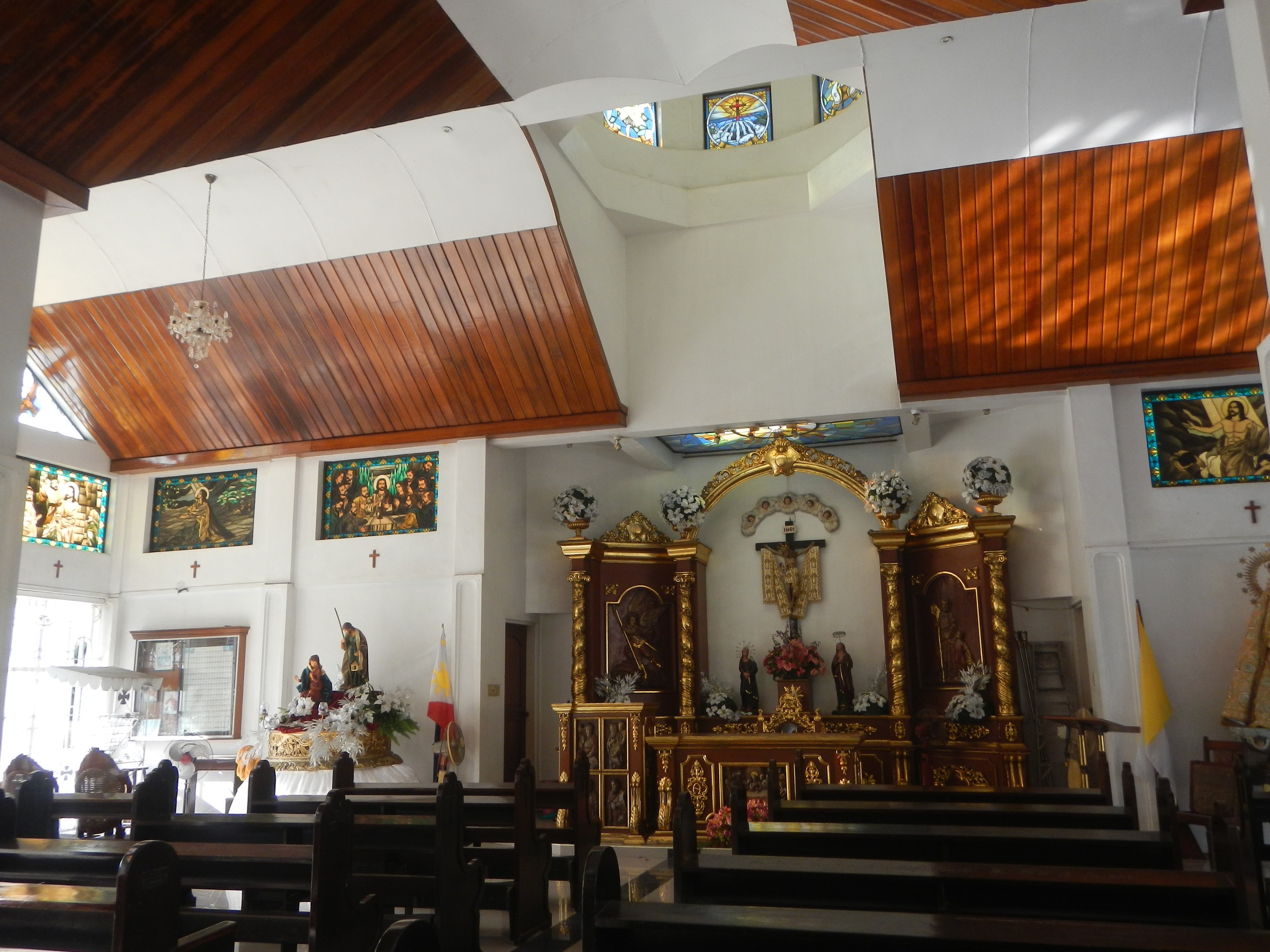 Innere einer Kirche mit Holzlehnen, Statuen, Blumenbouquets, Fahnen, Fotorahmen an den Wänden und Buntglasfenstern, Decke mit Lichtern geschmückt.