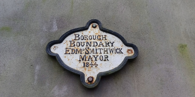 Metallplakette an einem Gebäude mit schwarzem Hintergrund und weißer fettgeschriebener Schrift, die "Borough Boundary Edm Smithwick Mayor" liest.