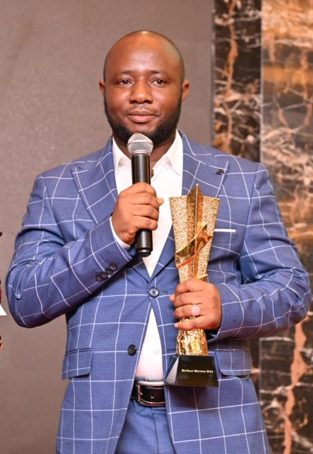 Ein Mann in einem blauen Anzug mit einem Mikrofon und einem Pokal, der selbstbewusst lächelt und vor einer Wand steht.