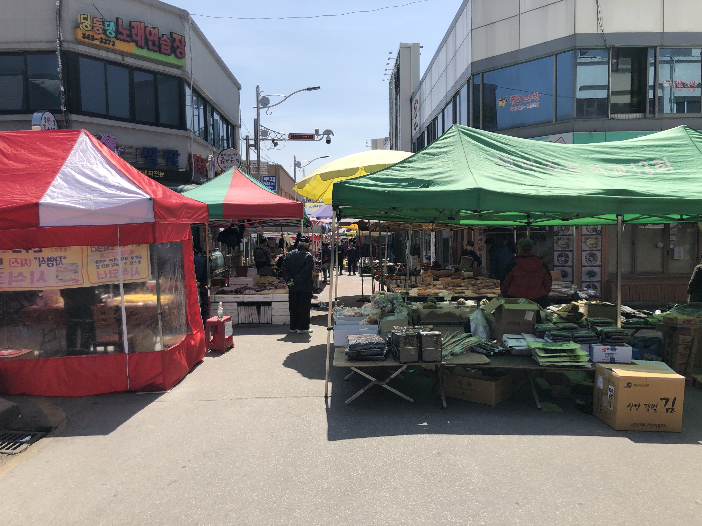 Ein belebter Straßenmarkt in Seoul, Südkorea, mit Zelten, Menschen und Tischen mit verschiedenen Artikeln entlang der Straße, vor Buildings mit Fenstern und Schildern unter einem sichtbaren Himmel.