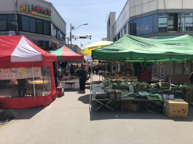 Ein belebter Straßenmarkt in Seoul, Südkorea, mit Zelten, Menschen und Tischen mit verschiedenen Artikeln entlang der Straße, vor Buildings mit Fenstern und Schildern unter einem sichtbaren Himmel.