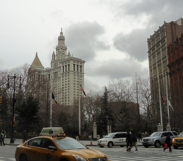 Gelbes Taxi fährt auf einer Stadtstraße mit hohen Gebäuden, Passanten auf dem Gehweg, Verkehrskegeln, Laternenmästen, Fahnenmästen, Bäumen und einem bewölktem Himmel.