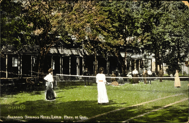 Schwarz-weißes Foto einer Gruppe von Menschen, die Tennis auf dem Rasen vor dem Abenakis Springs Hotel in Provo, Quebec, spielen, mit einem Netz, Stühlen, Bäumen und einem Gebäude im Hintergrund.