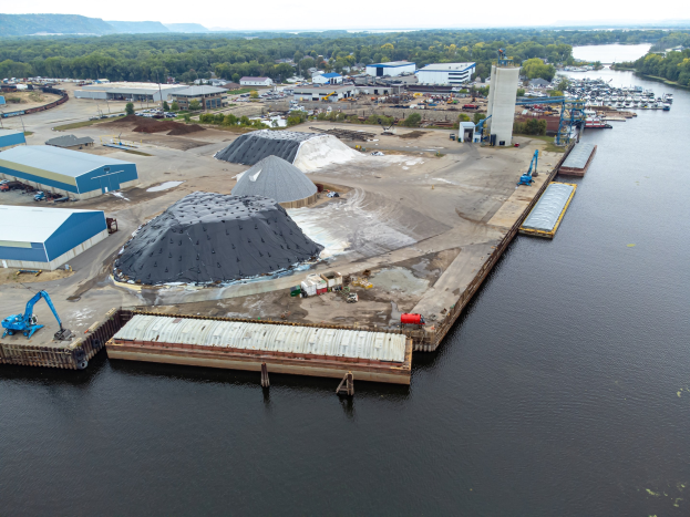 Luftaufnahme eines großen Gewässers mit einem zentralen Kai, umgeben von Gebäuden, Schuppen, Fahrzeugen und Kränen, vor Wald, Hügeln und einem klaren blauen Himmel.