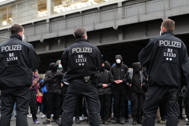 Polizeibeamte in schwarzer Uniform und Masken stehen vor einer Menge, mit einer Brücke und einem Gebäude im Hintergrund.