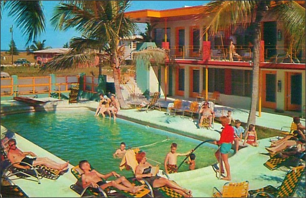 Ein Schwimmbad mit Menschen auf Stühlen im Vordergrund, Gebäuden und Bäumen im Hintergrund und sichtbaren Autos.