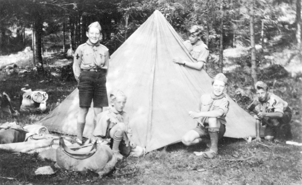 Schwarz-weiß-Foto von Pfadfindern in Uniform, die sich um ein Zelt in einem bewaldeten Gebiet versammelt haben, einige sitzen auf dem Boden, während andere stehen, mit verstreuten Taschen und Ausrüstung.