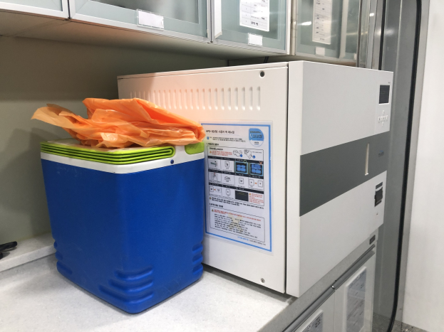 Blauer und weißer Kühler auf einer Theke neben einer Ultraschallreinigungsmaschine, mit einer Kunststoffabdeckung oben und anderen Gegenständen in der Nähe, unter Hängeschränken.
