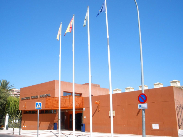 Großes Gebäude mit Flaggen vorne, als Hauptquartier der spanischen Nationalpolizei identifiziert, umgeben von Straßeninfrastruktur und Bäumen unter einem klaren blauen Himmel.