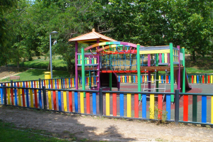 Ein Spielplatz mit einer bunten Spielstruktur in der Mitte, umgeben von einem Zaun, mit Pfosten, Laternen, Bänken, Gras, Pflanzen und Bäumen, unter einem klaren blauen Himmel.