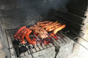 Grill mit Würstchen und Fleisch darauf, umgeben von Kohle und Feuer, im Freien auf Gras stehend.