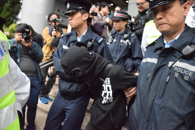 Eine Gruppe von Polizisten steht vor einer Menge von Menschen, einige tragen Mützen und halten Kameras, mit einem Gebäude mit Säulen und Deckenleuchten im Hintergrund und Bäumen links.