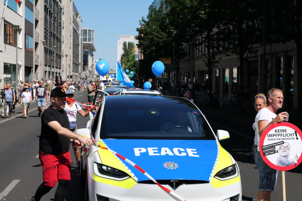 Eine Gruppe von Menschen neben einem Polizeiwagen mit blauen, gelben und roten Bändern und dem Wort "Frieden", mit Gebäuden, Bäumen, Fahnen und einem klaren Himmel im Hintergrund.