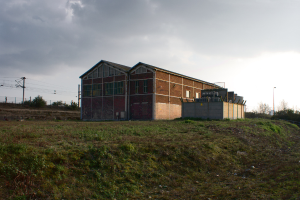Eine Szene mit Schuppen, Transformatoren, Strommasten mit Kabeln, Zäunen, einer Straßenlaterne, Bäumen und einem bewölkten Himmel.