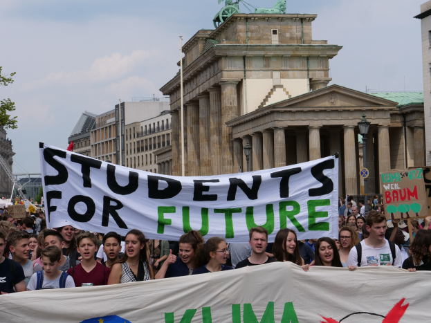 Gruppe von Schülern marschiert in Berlin mit einer bunten "Schüler für die Zukunft"-Fahne an Gebäuden, Bäumen und Himmel vorbei.