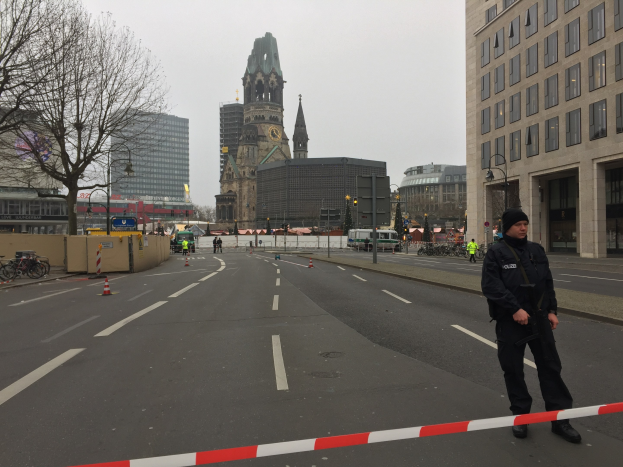Polizist in schwarzer Uniform und Mütze auf einer Berliner Straße umgeben von Absperrbaken, Fahrrädern, Laternenpfählen, Bäumen und Gebäuden mit einem Kirchturm und einem klaren blauen Himmel im Hintergrund.