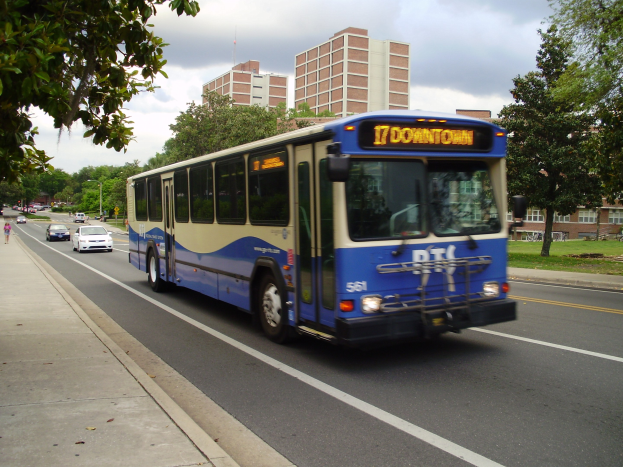 Blauer und weißer Shuttlebus fährt auf einer Straße mit hohen Gebäuden, Fußgängern auf dem Gehweg und Bäumen im Hintergrund unter einem klaren blauen Himmel.