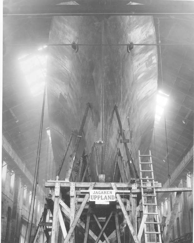 Großes Schwarz-Weiß-Foto eines Schiffes in einem Schiffbau, das eine Leiter, Holzpfosten, ein Schild, Metallrahmen, Deckenleuchten und Fenster zeigt.