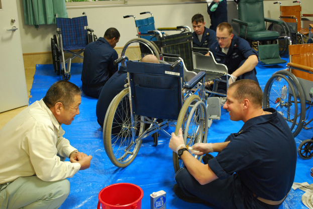 Eine Gruppe von Männern arbeitet an einem Rollstuhl in einem Raum mit mehreren Rollstühlen, einem Eimer und anderen Gegenständen auf dem Boden, mit einer Tür links und Fenstern mit Vorhängen im Hintergrund.