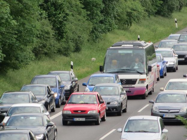 Verkehrsstau auf einer Autobahn mit mehreren Autos und einem Lieferwagen, mit Passagieren in Fahrzeugen, vor einem Hintergrund aus Bäumen und Gras.