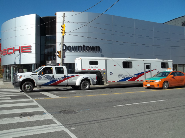 Ein Polizei-Lkw vor einem Gebäude mit Text, eine Ampel mit Stange und Drähten im Vordergrund und Bäume mit einem klaren blauen Himmel im Hintergrund.