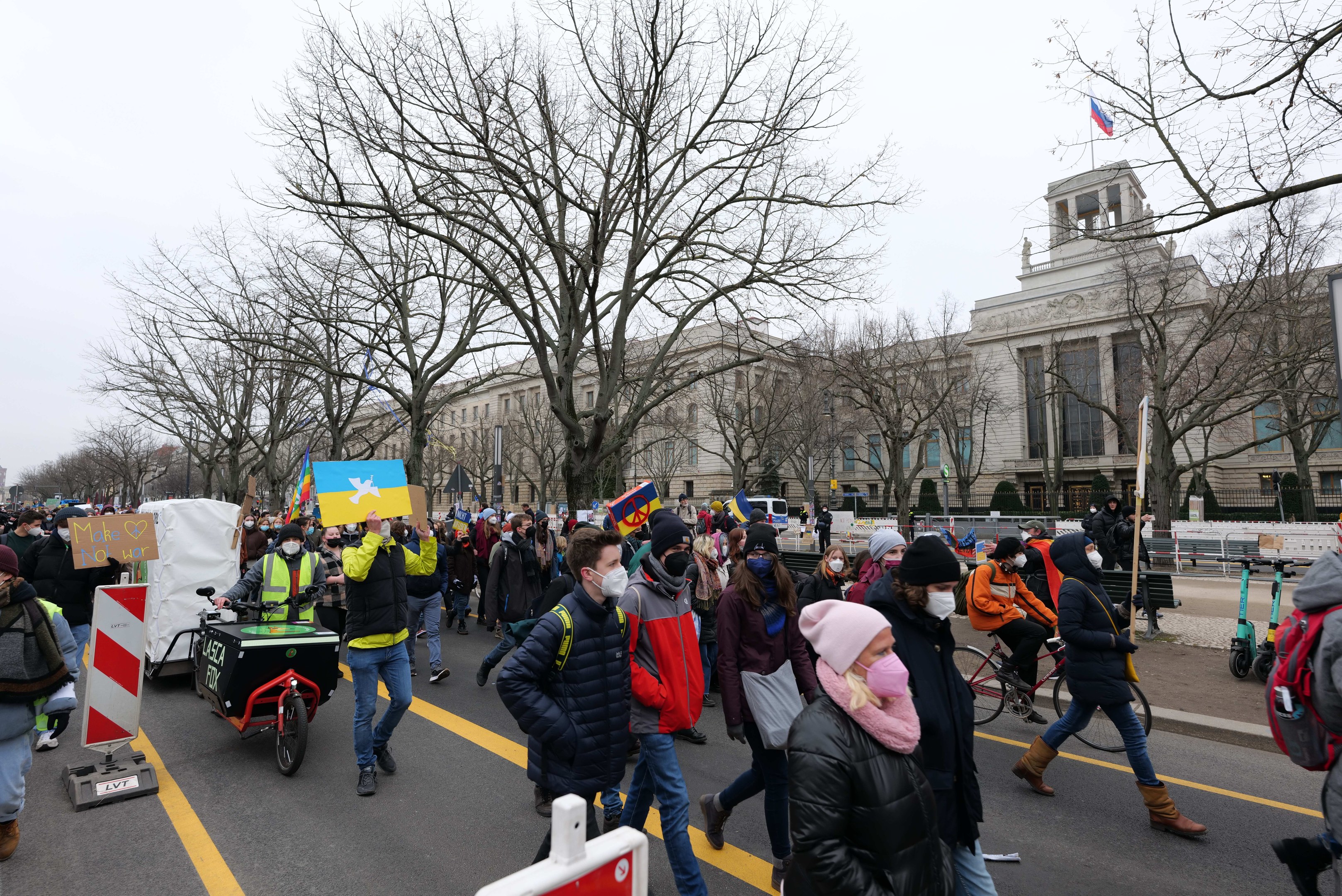 Eine große Gruppe von Menschen marschiert auf einer Stadtstraße in einem Protest, einige halten Schilder und fahren Fahrräder, mit Bäumen und einem Gebäude im Hintergrund unter einem klaren blauen Himmel.