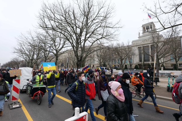 Eine große Gruppe von Menschen marschiert auf einer Stadtstraße in einem Protest, einige halten Schilder und fahren Fahrräder, mit Bäumen und einem Gebäude im Hintergrund unter einem klaren blauen Himmel.