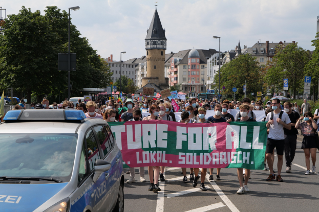 Eine große Gruppe von Menschen marschiert mit einem Banner mit der Aufschrift "Lokale Solidarität" eine Straße entlang, neben einem Streifenwagen geparkt, Bäume und Gebäude im Hintergrund unter einem bewölkten Himmel.