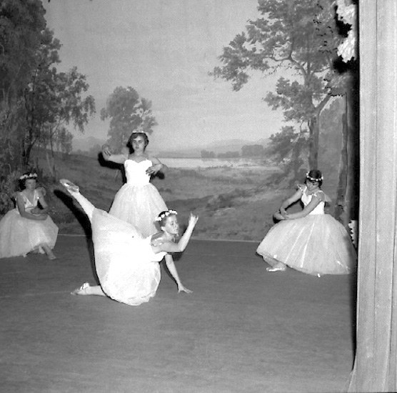 Vier junge Ballerinen in weißen Kleidern tanzen auf einer Bühne mit einer gemalten Kulisse aus Bäumen und Himmel in Schwarz-Weiß.