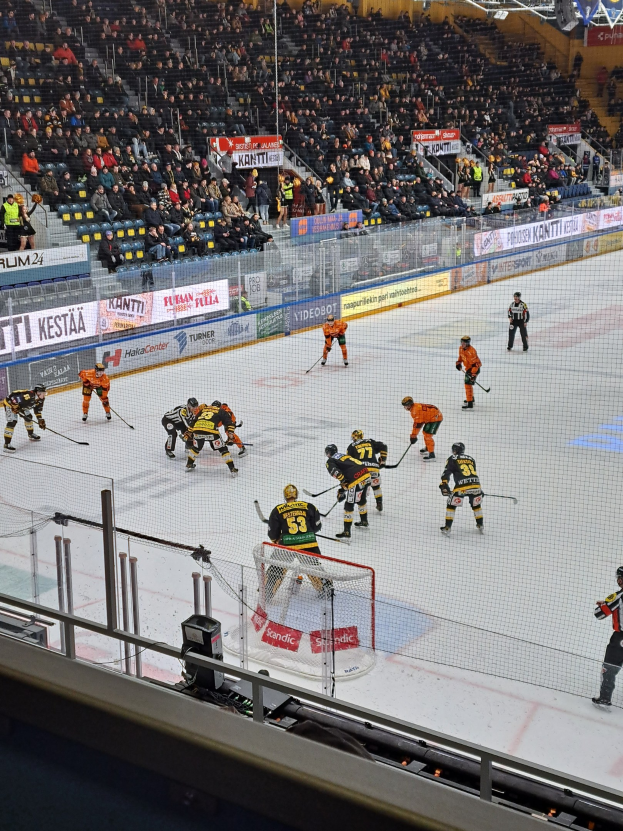 Eine Gruppe von Menschen, die Hockey auf einem Eisring mit einem Netz und einem Zaun spielen, Zuschauer sitzen im Hintergrund und Banner mit Text, unter Stadionbeleuchtung.