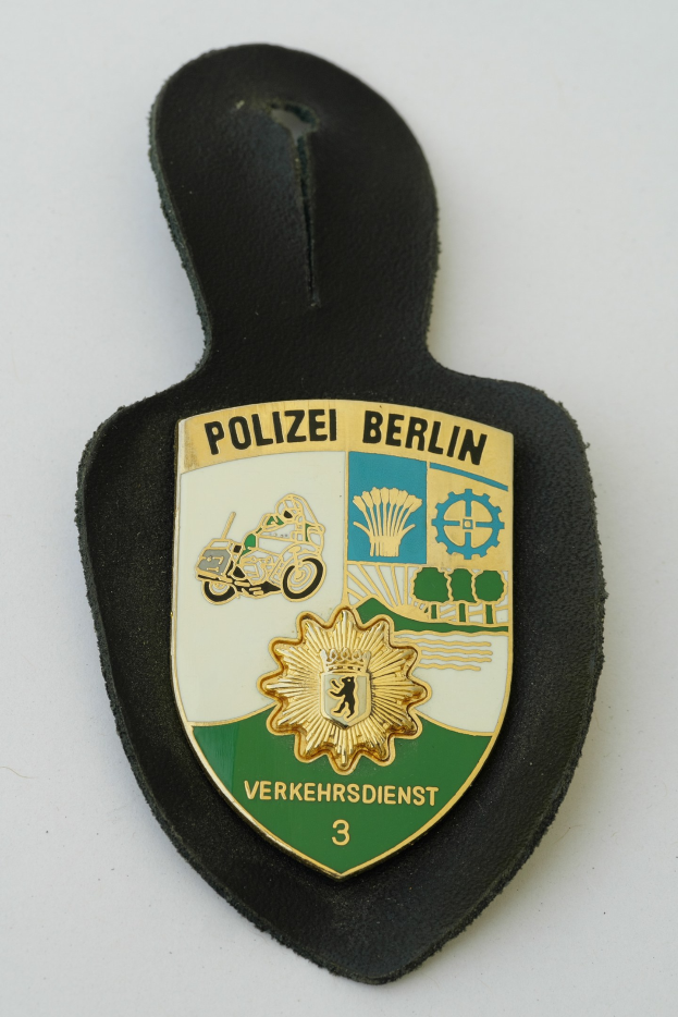 Ein schildförmiges Polizeibadge mit "Polizei Berlin" in schwarzer fettgeschriebener Schrift auf einem blauen Hintergrund mit einem weißen Stern und Rahmen, das auf einer Oberfläche ruht.