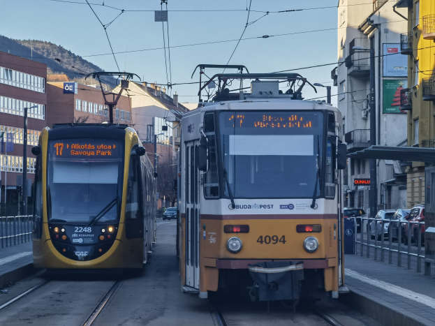 Zwei gelbe und weiße Straßenbahnen auf einer Stadtstraße mit hohen Gebäuden, Fahrzeugen, Geländern, Bäumen, einem Hügel und dem Himmel im Hintergrund.