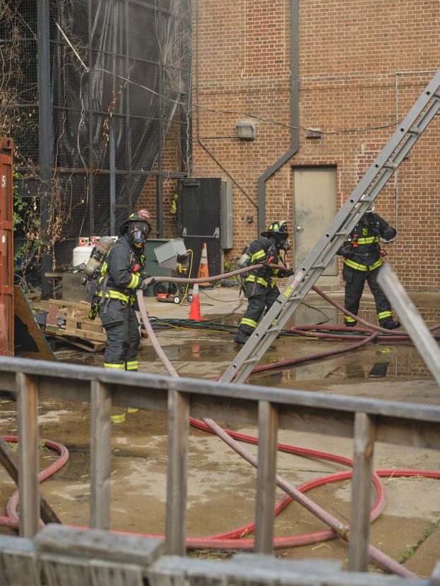Eine Gruppe von Feuerwehrleuten in Helmen, die Rohre halten, arbeiten daran, ein Gebäude Feuer zu löschen, wobei verschiedene Ausrüstungen und Strukturen in der Szene zu sehen sind.