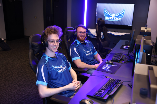 Zwei Männer mit Headsets lächeln, während sie an einem Schreibtisch mit Computermonitoren, Tastaturen und anderem Equipment sitzen und scheinbar ein Videospiel in einem Raum mit einem Bildschirm und Lichtern im Hintergrund spielen.