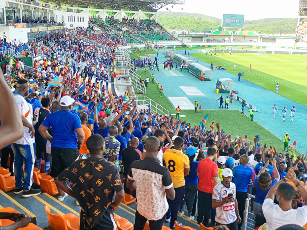 Eine große Menschenmenge versammelt sich vor einem eingezäunten Fußballfeld, wobei einige auf Stühlen sitzen und andere stehen, während Hügel und ein klarer blauer Himmel im Hintergrund zu sehen sind.