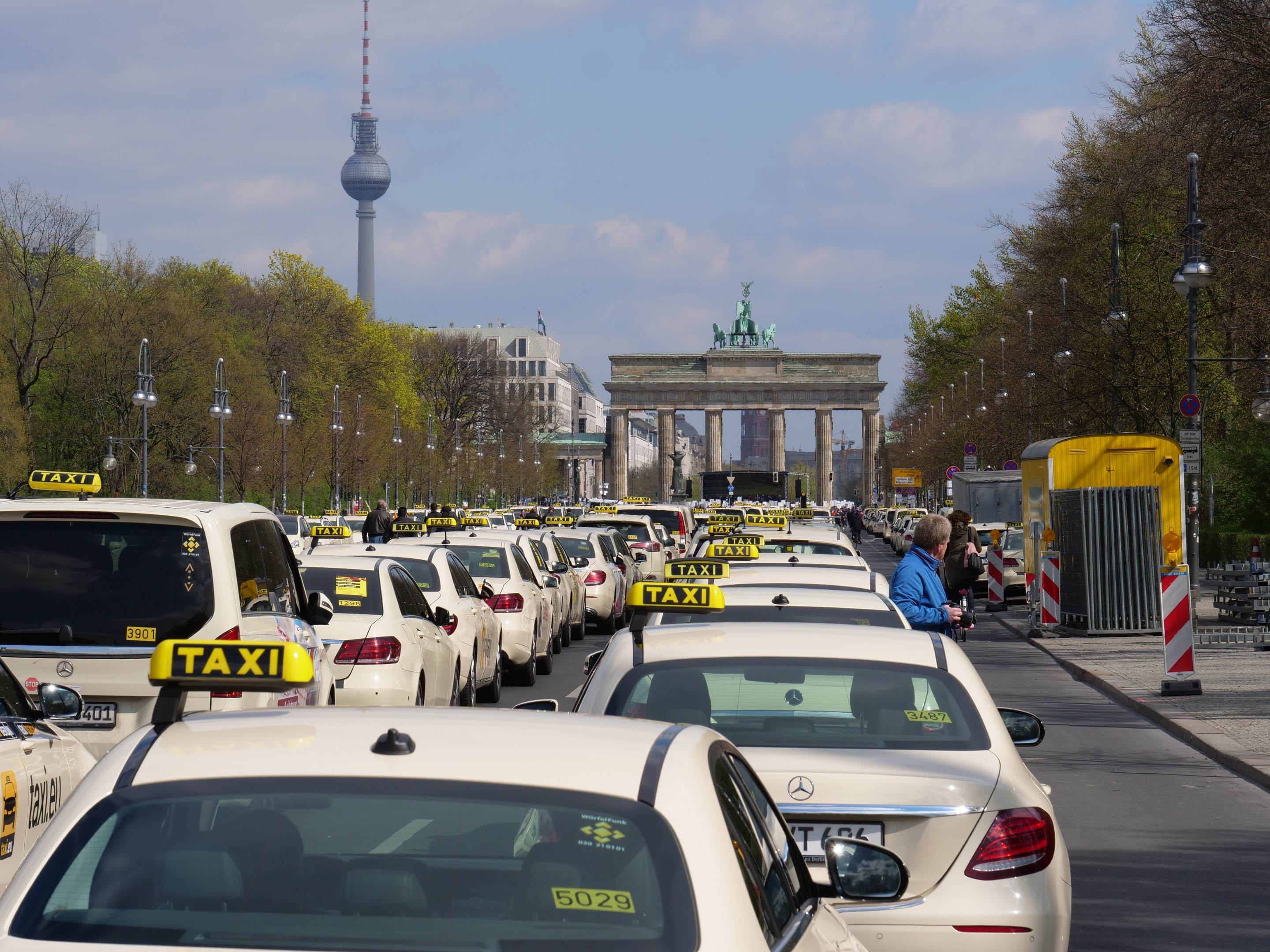 Eine belebte Straße in Berlin mit zahlreichen parkenden Taxis, Fußgängern auf dem Gehweg, gesäumt von Laternenpfählen und Bäumen, Gebäuden im Hintergrund, einem fernen Bogen mit Statuen und Turm und einem bewölkten Himmel.