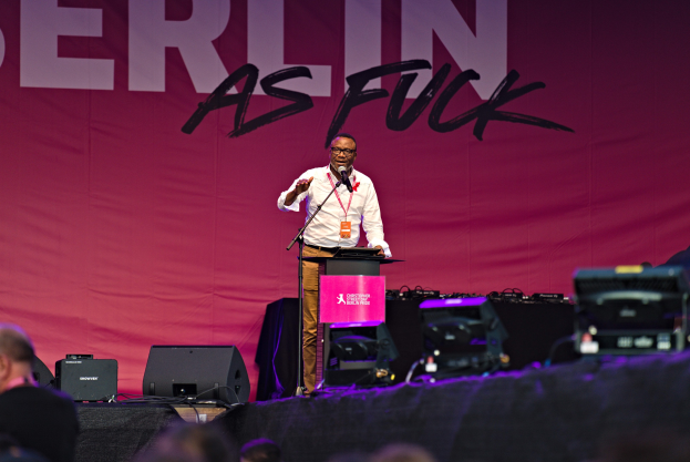 Mann in weißem Hemd und braunen Hosen, der in ein Mikrofon spricht, vor einem pinken Banner 'Berlin as Fuck' auf einer Bühne mit Publikum