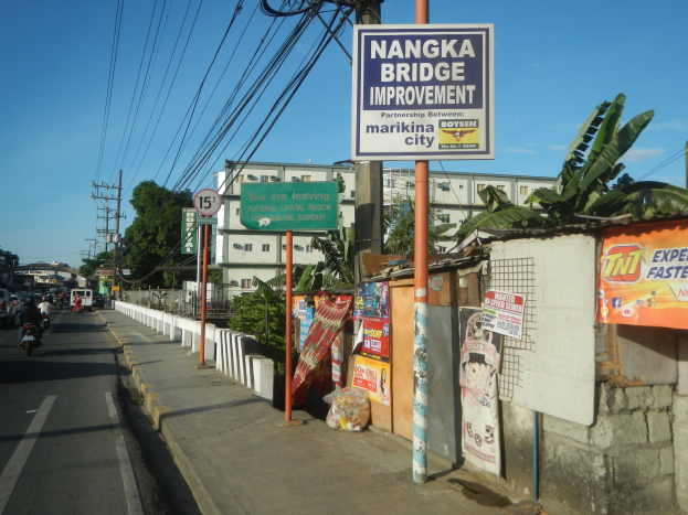 Straße mit Fahrzeugen, ein Bürgersteig-Schild mit der Aufschrift "Nangka Bridge Improvement", Gebäude, Bäume, Strommasten mit Drähten und ein bewölkter Himmel im Hintergrund.