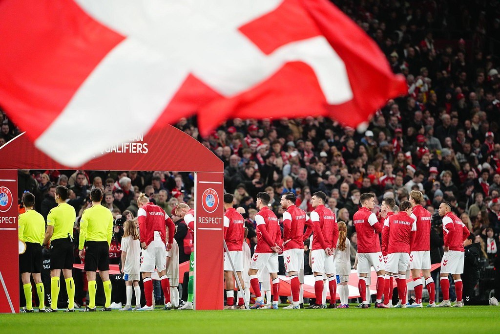 Eine Gruppe von Menschen auf einem Fußballfeld mit einer roten und weißen Flagge im Vordergrund, einem Bogen mit der Aufschrift "Bayern München vs. Bayern München Betting Tips & Preview" im Hintergrund und einer großen Menge im Stadion.