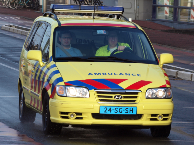 Ein gelbes Rettungsfahrzeug mit zwei sichtbaren Insassen fährt an parkenden Fahrrädern auf einem Gehweg vorbei, mit einem Glasfenstergebäude im Hintergrund.