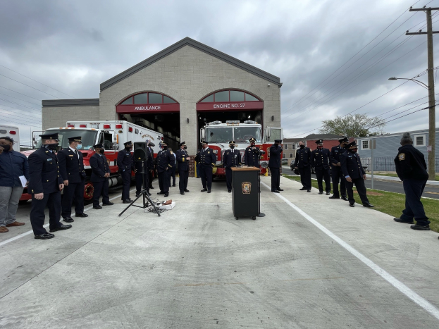 Gruppe von Menschen in Kappen neben einem Löschfahrzeug auf einem Podium während einer Feuerwache-Einweihungszeremonie, mit Gebäuden, Bäumen, Strommasten und Gras im Hintergrund unter einem bewölkten Himmel.