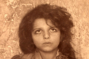 Ein Schwarz-Weiß-Foto eines jungen Mädchens mit langen lockigen Haaren, das vor einer Wand steht, mit einem Wasserzeichen in der rechten unteren Ecke.