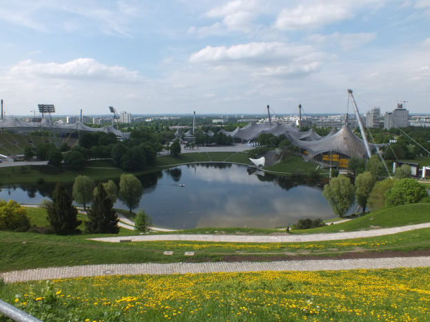 Ein Blick auf das Olympiastadion von einem Hügel aus, mit einem Teich im Vordergrund, umgeben von Grün und gelben Blumen, und Gebäuden im Hintergrund unter einem klaren blauen Himmel.