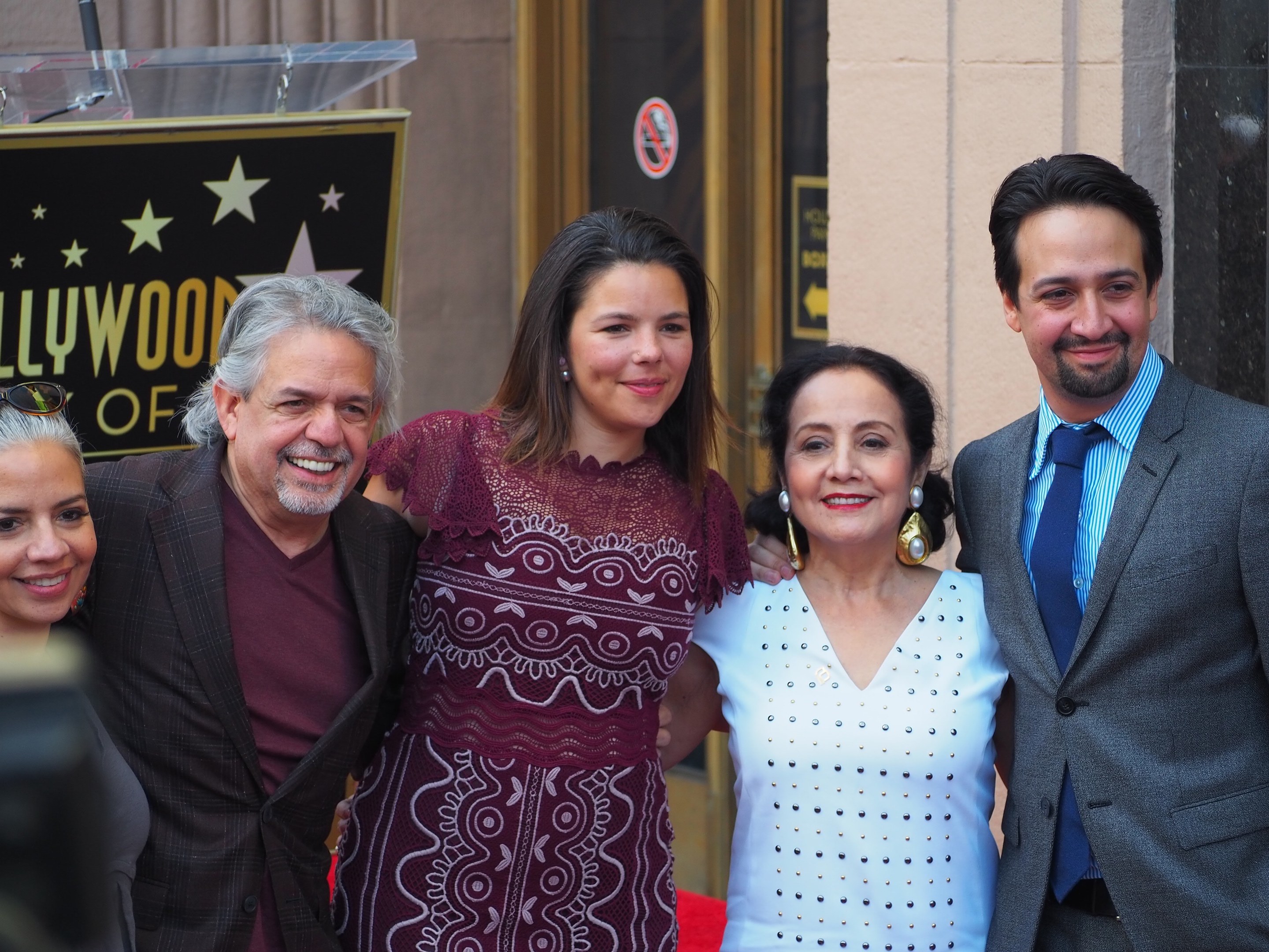 Gruppe von Menschen, die auf einer Preisverleihung auf dem Roten Teppich posen, mit einer Kamera auf der linken Seite, einer Tafel mit Text, einem Glastisch und einer Wand im Hintergrund.