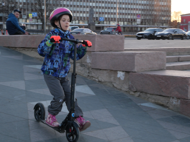 Ein junger Junge in Helm und Handschuhen fährt einen Roller eine Treppe hinunter, vorbei an Fahrzeugen, Menschen, Bäumen, Pfosten, Brettern, Gebäuden und einem klaren blauen Himmel.
